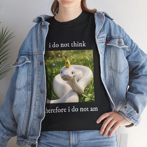 May include: Black t-shirt with a graphic of a white snake wearing a banana hat, set in grass. The text "i do not think" is above the image, and "therefore i do not am" is below. The shirt is worn by a person.