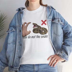 May include: White t-shirt with a graphic of a hand, a snake, and a red X. The text on the shirt reads "Please do not the snake". The snake is brown and tan with its mouth open.