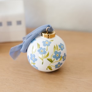 May include: A white ceramic Christmas ornament with a gold cap and a light blue ribbon. The ornament is decorated with hand-painted blue forget-me-not flowers and green leaves. Decorative holiday decor.