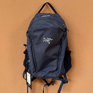 Puede incluir: Una mochila azul marino con asa y correas negras. La mochila tiene un logotipo blanco y la palabra "ARCTERYX" impresa en la parte superior. Cuenta con múltiples compartimentos con cremallera y correas ajustables.