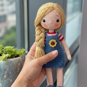 May include: A handmade crochet doll with blonde yarn hair styled in a side braid. The doll wears a blue overall dress featuring a small yellow sunflower appliqué on the chest, layered over a red and white striped short-sleeve shirt and blue shoes.