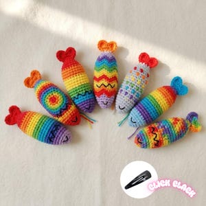 May include: Seven small, stuffed, crocheted fish ornaments in various rainbow color patterns. Each fish features a distinct tail shape and embroidered facial details. A black metal hair clip is shown in a circular inset with the text CLICK CLACK.
