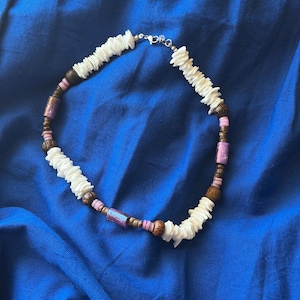 May include: A beaded necklace featuring white shell segments, dark brown wooden beads, small light pink disc beads, and cylindrical purple metallic beads. The necklace is secured with a silver-toned metal lobster claw clasp and jump ring.