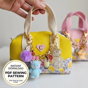 May include: A yellow handbag with cream-colored handles and gold hardware, adorned with charms including a blue teddy bear, a purple pom-pom, and a seashell. The bag has a floral patterned base and a pink heart with the letter "C". The image includes text that says "Instant Download PDF Sewing Pattern".