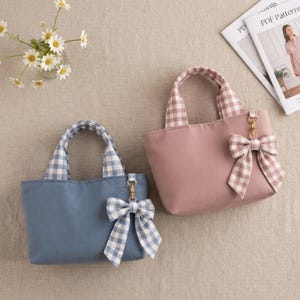 May include: Two small tote bags with handles and a decorative bow attached to a gold-tone clasp. One bag is dusty blue with blue and white gingham handles and a matching bow. The other bag is dusty pink with pink and white gingham handles and a matching bow.