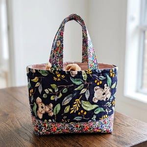 May include: A quilted fabric tote bag featuring a dark navy blue background with a pattern of koalas, green leaves, and yellow berries. The base and handles are accented with a vibrant multi-colored floral print, and the interior is lined in pink.