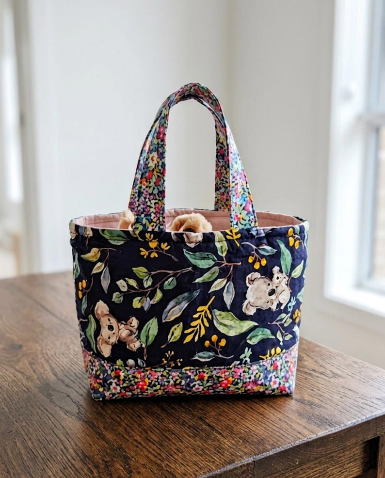 May include: A quilted fabric tote bag featuring a dark navy blue background with a pattern of koalas, green leaves, and yellow berries. The base and handles are accented with a vibrant multi-colored floral print, and the interior is lined in pink.