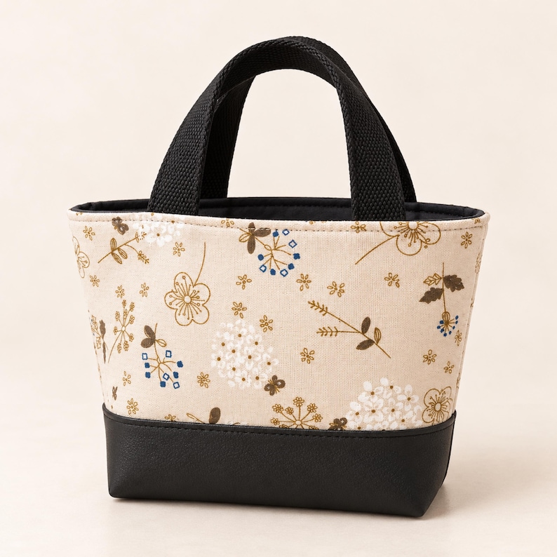 May include: A small tote bag featuring a beige fabric body with a printed pattern of brown, white, and blue flowers and foliage. The bag has a solid black base and two black woven fabric handles attached to the top edge of the bag.