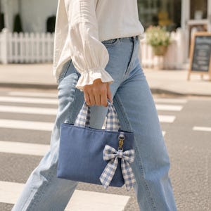 A small navy blue canvas handbag featuring two short handles with a light blue and white gingham check pattern. A matching gingham bow is attached to the front of the bag with a small gold-toned metal lobster clasp hardware.