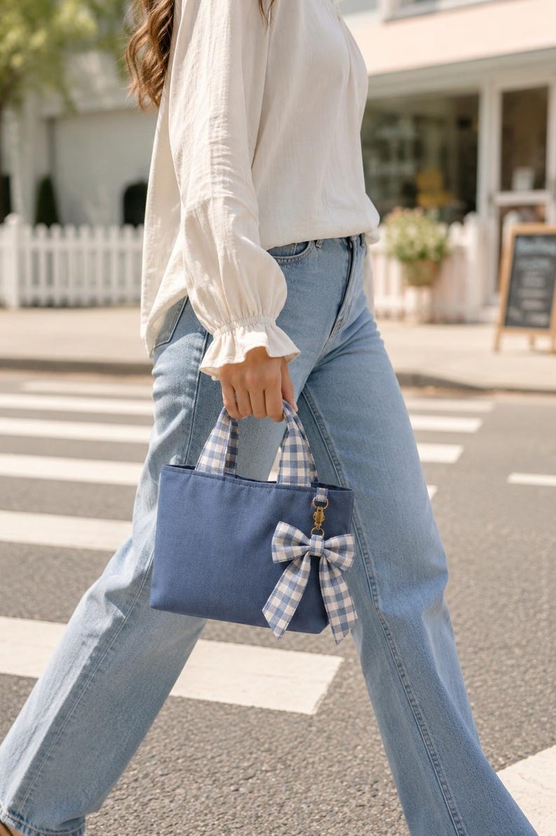 A small navy blue canvas handbag featuring two short handles with a light blue and white gingham check pattern. A matching gingham bow is attached to the front of the bag with a small gold-toned metal lobster clasp hardware.