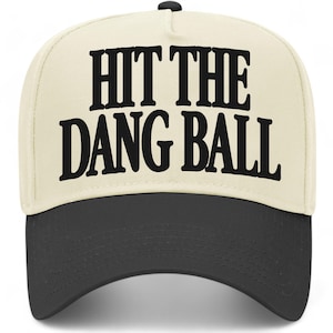 May include: A baseball cap with a cream-colored crown and a dark gray brim. The cap features the text "HIT THE DANG BALL" in bold, black letters across the front. The cap has a black button on top.