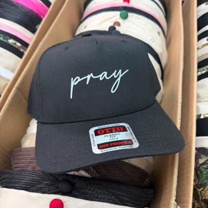 Op de afbeelding: Zwarte baseball cap met het woord "pray" in witte letters op de voorkant. De cap heeft een gebogen klep en een label met de tekst "OTTO Classic Fit". Andere caps zijn zichtbaar op de achtergrond.