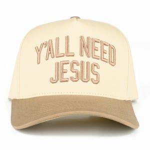 May include: A cream-colored baseball cap with a brown brim. The phrase "Y'ALL NEED JESUS" is embroidered in a light brown color across the front of the cap. The cap is a classic six-panel design.