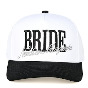May include: White baseball cap with a black brim. The word "BRIDE" is embroidered in bold black letters, with "Needs a Margarita" embroidered below in silver script. A fun accessory for a bachelorette party.
