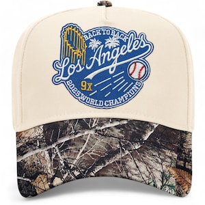 May include: Cream-colored baseball cap with a camouflage brim. The cap features a blue and white emblem with the text "Back to Back Los Angeles 2025 World Champions" and a baseball graphic.