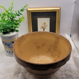 May include: A large, brown ceramic bowl with a textured interior and decorative bands around the exterior. The bowl sits on a white lace tablecloth. A framed picture and a small potted plant are in the background.