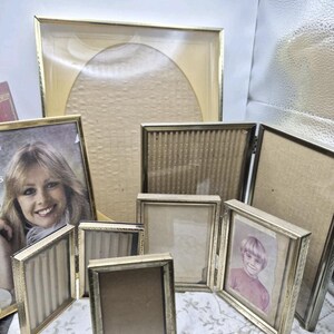 May include: A collection of seven gold-toned metal picture frames of varying sizes and shapes. The set includes rectangular and oval-shaped frames, some featuring hinged designs. Two frames contain vintage-style portrait photographs.