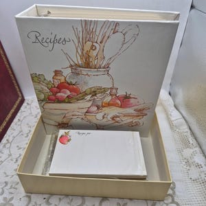 May include: A white three-ring binder with the word Recipes printed in script, featuring a kitchen illustration of vegetables and utensils. It includes a matching cream-colored box containing a stack of white recipe cards with a strawberry graphic.
