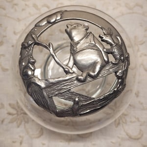 May include: A clear glass bowl featuring a circular pewter lid. The lid displays a relief sculpture of a bear sitting on a branch with a small pig-like creature, a rabbit, and a bird. The metal surface has a matte, silver-toned finish.