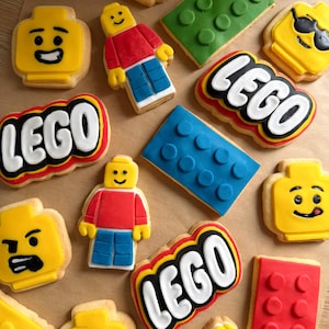 May include: A collection of sugar cookies shaped like yellow minifigure heads with various facial expressions, red and blue minifigure bodies, rectangular building bricks in red, green, and blue, and cookies featuring the white LEGO logo on red.
