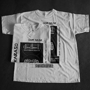 May include: Two white t-shirts with black graphic designs. One shirt is folded with a tag that reads "LARGE". The designs include the text "OOM GEAR", "KARASO", and technical diagrams. The shirts are on a dark surface.
