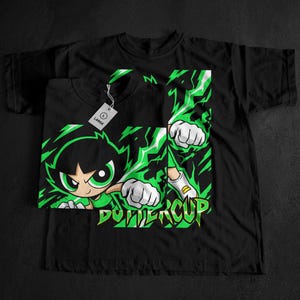 Puede incluir: Camisetas negras con un gráfico de Buttercup de Las Supernenas. El diseño presenta a Buttercup en una pose dinámica con detalles en verde y blanco, y la palabra "Buttercup" en verde. Una etiqueta "Large" está adjunta.