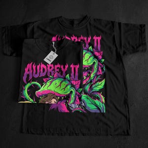 May include: Black t-shirts featuring a vibrant graphic of Audrey II, a carnivorous plant, with the text "Audrey II" in pink and purple. The design includes green and purple plant details. One shirt has a "LARGE" tag.