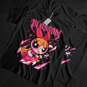 Op de afbeelding: Zwarte t-shirts met een afbeelding van Blossom van The Powerpuff Girls. Het ontwerp bevat de afbeelding van Blossom, het woord "BLOSSOM" in roze en roze en witte accenten. Een label luidt "LARGE".
