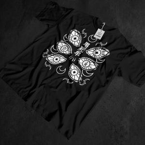 Może przedstawiać: Czarny t-shirt z białym graficznym wzorem stylizowanych oczu ułożonych w okrąg. Projekt zawiera japoński tekst i motywy półksiężyca. Do koszulki przymocowana jest biała metka z napisem "LARGE".