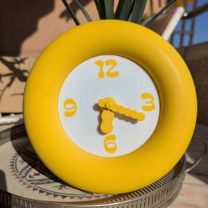 Puede incluir: Un reloj analógico redondo con un marco grueso de color amarillo brillante y una esfera blanca. El reloj cuenta con manecillas de hora y minutos amarillas con forma ondulada, junto con números grandes, amarillos y redondeados para 12, 3, 6 y 9.