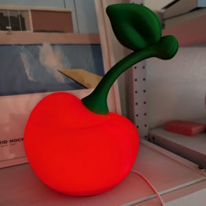 May include: A cherry-shaped lamp with a vibrant, glowing red spherical body and a solid green stem featuring a single leaf. The lamp is connected to a thin white power cord that extends from the base of the red fruit shape.