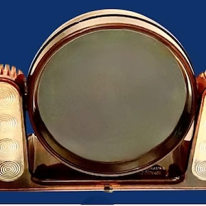 May include: Vintage Art Deco vanity mirror with a circular frame and two side light fixtures. The mirror has a bronze finish and a dark gray reflective surface. The side lights have a series of concentric circles.