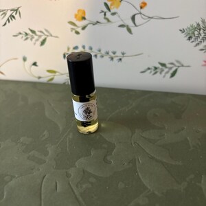 May include: A small glass roller bottle with a black cap, filled with a yellow liquid. The bottle has a white label with text and a floral design. The background features a floral pattern with pink and yellow flowers.
