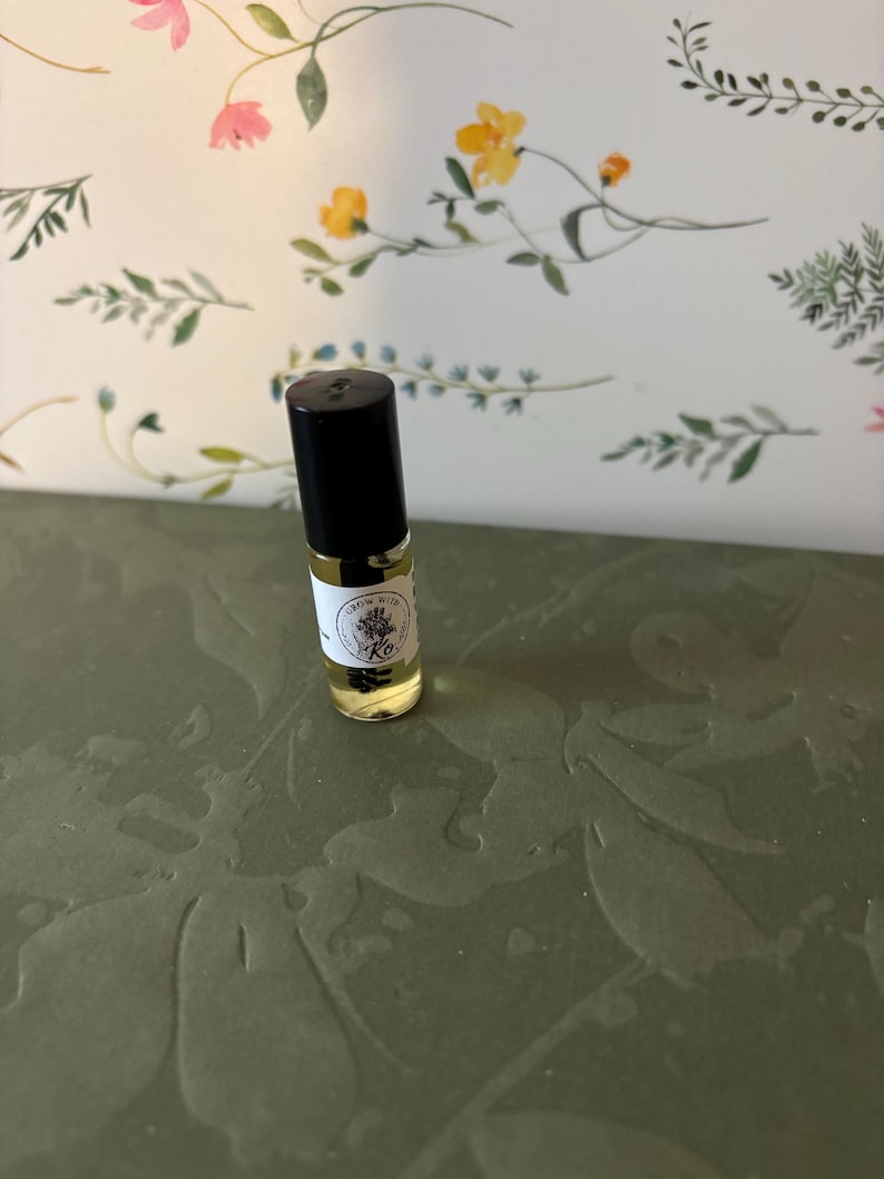 May include: A small glass roller bottle with a black cap, filled with a yellow liquid. The bottle has a white label with text and a floral design. The background features a floral pattern with pink and yellow flowers.