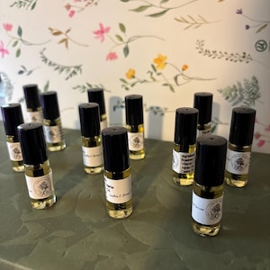 May include: A collection of small, clear glass essential oil bottles with black caps. Each bottle has a white label with text and a floral design. The bottles are arranged on a green surface, with a floral patterned background.