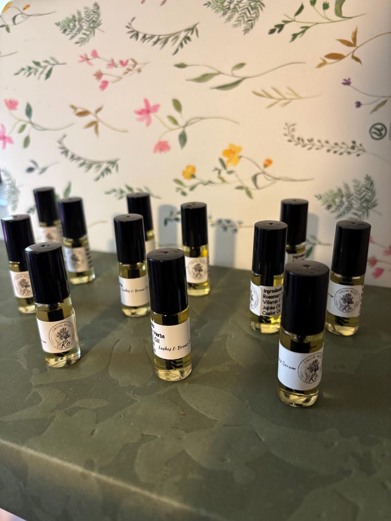 May include: A collection of small, clear glass essential oil bottles with black caps. Each bottle has a white label with text and a floral design. The bottles are arranged on a green surface, with a floral patterned background.