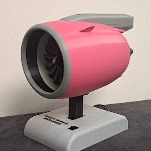 Può includere: Un modello di motore di aereo rosa e grigio, con un supporto nero e una base grigia. Il motore ha una ventola all'interno e la base ha il testo "Josh & Walker Studios" e un interruttore. Il modello è su una superficie grigio scuro.