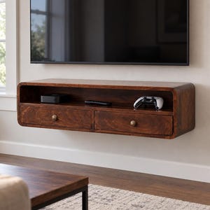 May include: A wall-mounted floating media console crafted from dark brown wood with rounded edges. The unit features an open top shelf and two lower drawers, each equipped with a single small, round, metallic knob for easy access and storage.