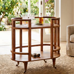 May include: An oval wooden bar cart featuring two tiers with a natural finish. The top tier has a woven cane surface, while the bottom tier is solid wood. The cart is supported by four legs with small metal casters for mobility.