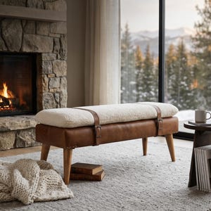May include: A brown leather bench with a cream-colored, fluffy cushion. The bench has wooden legs and is secured with leather straps. The bench is approximately 48 inches long.