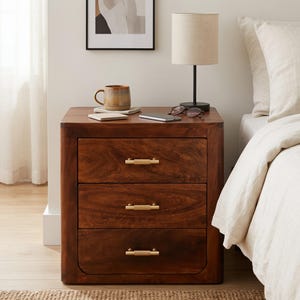 May include: A dark brown wooden nightstand with three drawers, each featuring a horizontal brushed gold handle. The nightstand has rounded corners and a smooth finish, providing a sturdy surface for a ceramic mug, a smartphone, and eyeglasses.