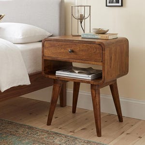 May include: A wooden nightstand with a rounded rectangular frame, a single drawer featuring a small dark knob, and an open storage shelf below. The piece stands on four tapered legs and holds a glass lamp, a small bowl, and a book titled Mid-Century Design.