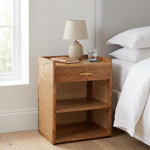 May include: A wooden nightstand featuring a single drawer with a gold-toned handle, two open storage shelves, and side panels crafted from woven cane webbing. The nightstand is topped with a beige table lamp and a clear glass of water.