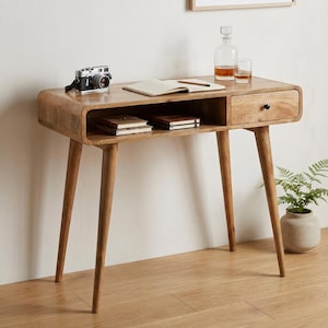 May include: A light brown wooden desk with rounded edges, four tapered legs, an open storage shelf, and a single drawer with a small black knob. The desk surface holds a silver camera, an open notebook, a glass decanter, and a small glass.