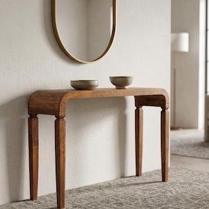 May include: A medium-brown wooden console table with a smooth, curved top edge and four straight, tapered legs. Two small, round, neutral-toned bowls are placed side-by-side on the flat surface of the table, centered between the legs.