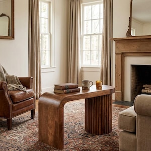 May include: A wooden coffee table with a curved design and a ribbed base. The table is a warm brown color and has a smooth surface. Two books, a mug, and a candle sit on top of the table. The table is in a living room setting.