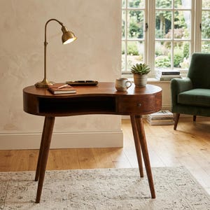 May include: A mid-century modern desk with a curved design and three angled legs. The desk is made of dark wood and features a drawer and a cutout for legroom. A brass desk lamp, a small succulent, and a mug are on the desk.