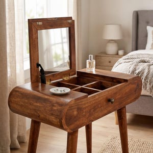 May include: A brown wooden vanity table with four tapered legs, featuring a hinged top that opens to reveal a mirror and four storage compartments. A small white dish with jewelry and a clear glass perfume bottle sit on the surface.