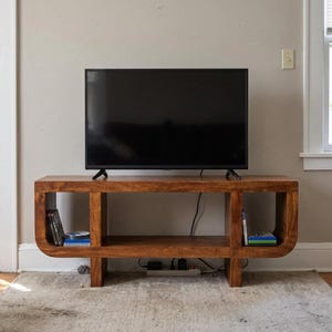 May include: A rectangular wooden media console with a warm brown finish and rounded edges, featuring two open side compartments and a central open shelf. A flat-screen television sits on top, and small stacks of media cases are stored on the sides.