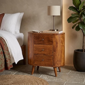 May include: A three-drawer wooden nightstand with a warm brown finish. The nightstand has a rounded front and tapered legs. A small lamp and a book titled "The Wes Anderson Collection" sit on top.